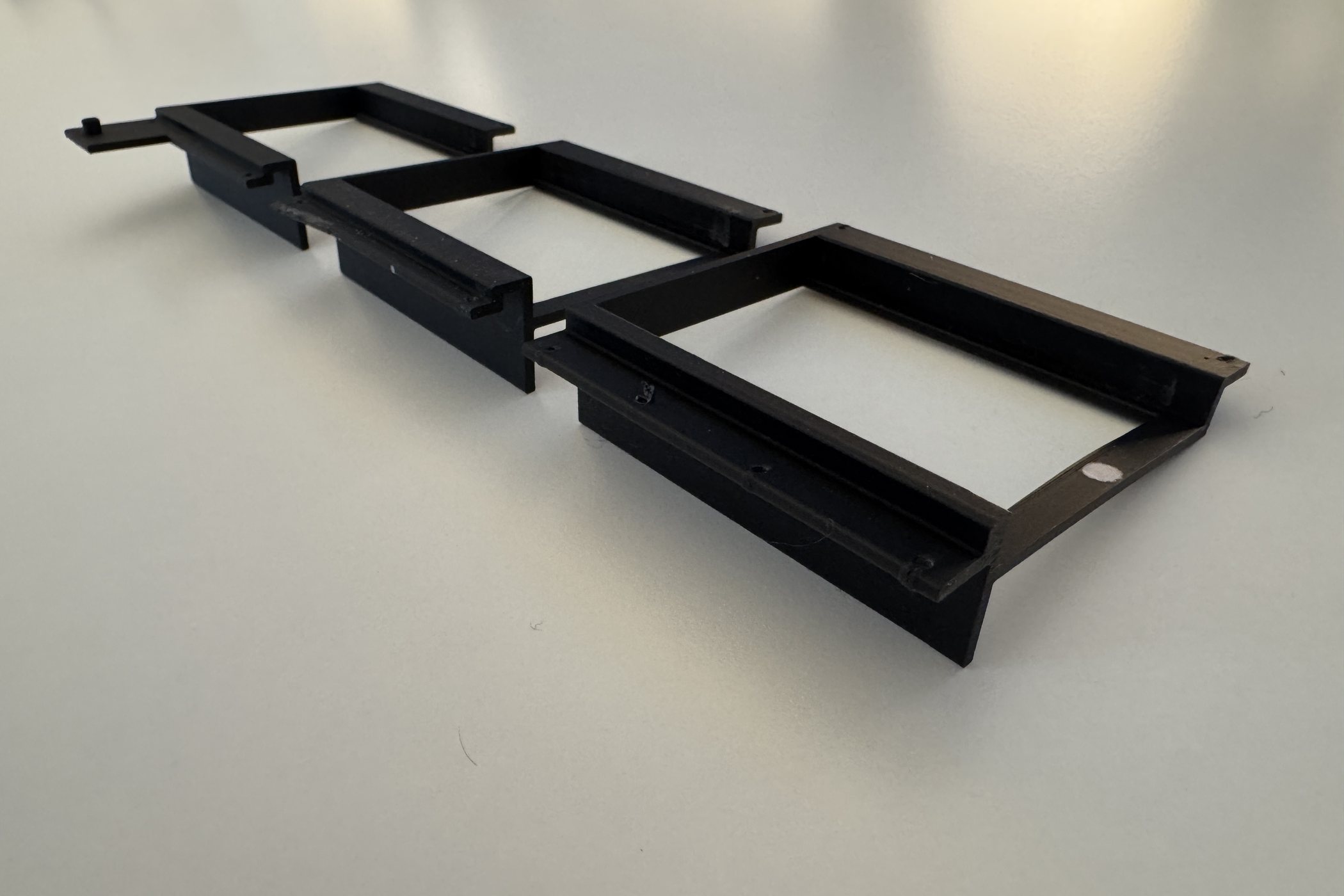 Three black 3D-printed display brackets photographed on a white surface, lined up in an overlapping row